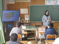 国際交流員の女性が黒板の前にたち、モニターを使いながら子供達に説明をしている写真