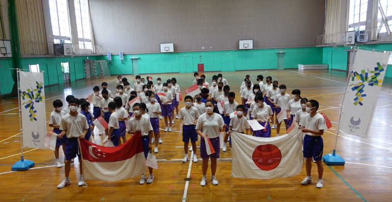 体育館で日本とシンガポールの国旗を持ち整列している児童の写真