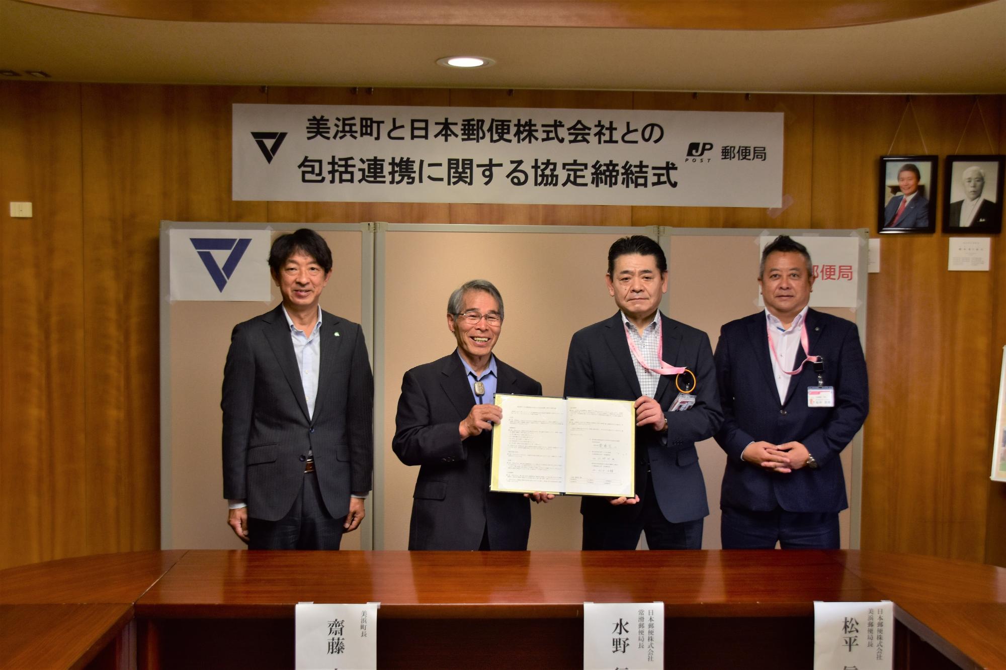 4人の関係者が立ち、その中央に町長 齋藤宏一氏と常滑郵便局長 水野行雄氏が協定書を持ち記念撮影している写真