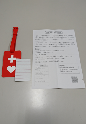 赤地に白で十字とハートマークが描かれたヘルプマークと「ヘルプマークについて」と書かれた紙が並べて置かれている写真