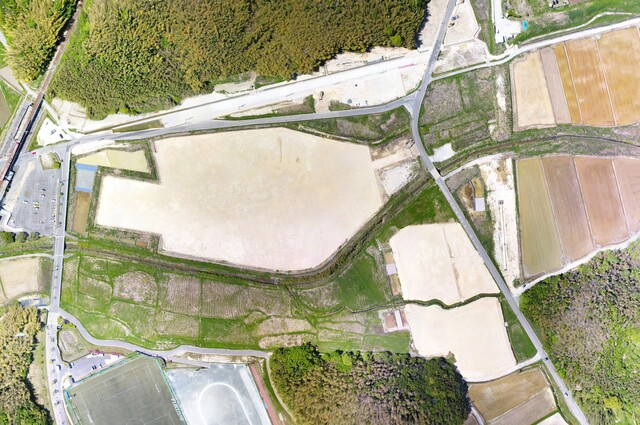 鬱蒼と茂った森と運動公園予定地の間を通る町道の工事が進んでいる様子を令和3年(2021)年4月上空から撮影した写真