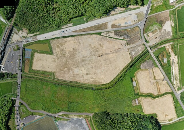 鬱蒼と茂った森と運動公園予定地の間を通る町道と、沈砂池と調整池の基盤設備の工事が進んでいる様子を令和3年(2021)年7月上空から撮影した写真