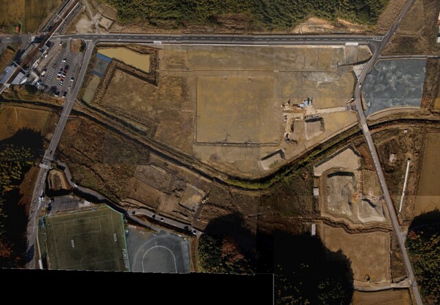 周囲を鬱蒼と茂った森が点在する建設予定地で調整池の設置工事が終了し、観覧スタンドの基盤設備工事が行われている様子を令和3年(2021)年12月上空から撮影した写真