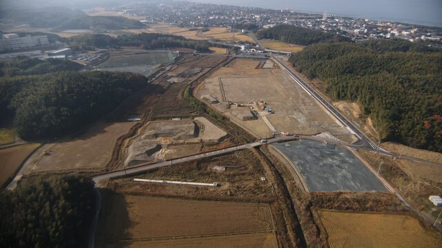 周囲に民家や鬱蒼と茂った森が点在する陸上競技場の建設予定地で、調整池の設置工事が完了し、観覧スタンドの基盤設備工事が進められている様子を令和3年(2021)年12月斜め上空から撮影した写真