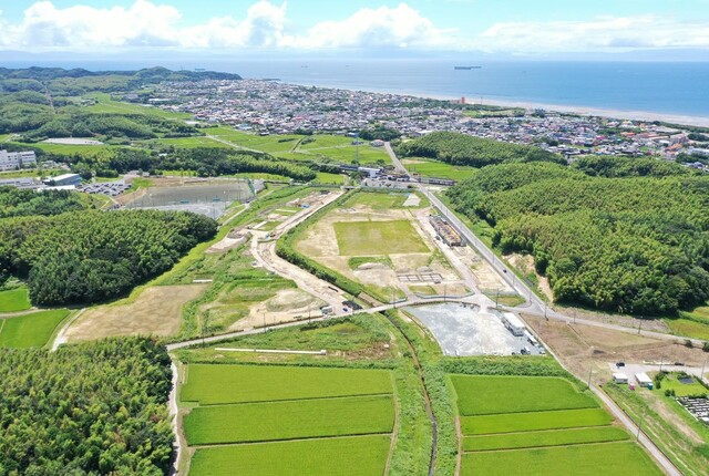 周囲に民家や鬱蒼と茂った森が点在する陸上競技場の建設予定地で、観覧スタンドと器具庫の工事が進められている様子を令和4年(2022)年7月斜め上空から撮影した写真
