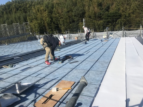 4名の作業員が陸上競技場観覧スタンド屋根部分に立ち、屋根材取付工事を行っている様子の写真