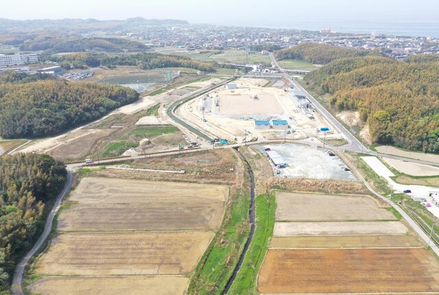 周囲に民家や鬱蒼と茂った森が点在する陸上競技場の建設予定地で工事が進められている様子を令和5年(2023)年4月斜め上空から撮影した写真