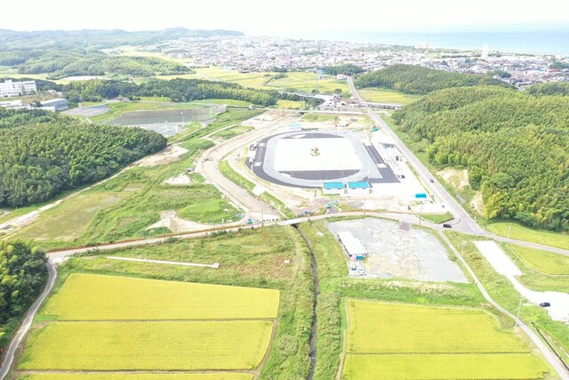 周囲に民家や鬱蒼と茂った森が点在する陸上競技場の建設予定地で工事が進められている様子を令和5年(2023)年8月斜め上空から撮影した写真