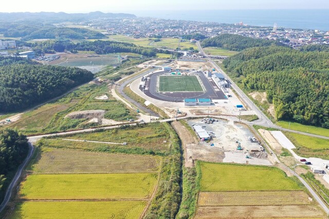 周囲に民家や鬱蒼と茂った森が点在する陸上競技場の建設予定地で工事が進められている様子を令和5年(2023)年10月斜め上空から撮影した写真