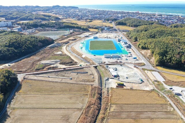 周囲に民家や鬱蒼と茂った森が点在する陸上競技場の建設予定地で工事が進められている様子を令和5年(2023)年12月斜め上空から撮影した写真