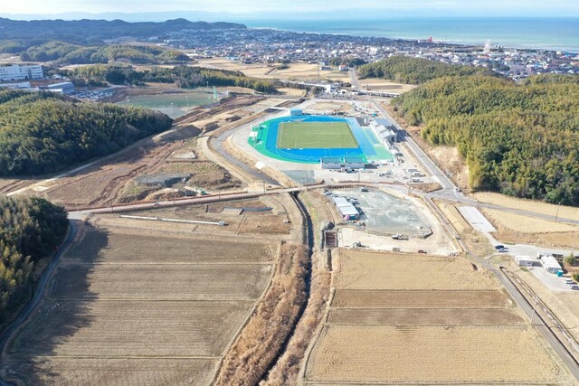 周囲に民家や鬱蒼と茂った森が点在する陸上競技場の建設予定地で工事が進められている様子を令和6年(2024)年1月斜め上空から撮影した写真