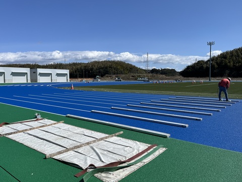 ヘルメットを被り防寒着を着用した作業員が、左側に白い器具庫が建っているのが見える場所のトラック部分のラインマーキング工事を行っている様子の写真