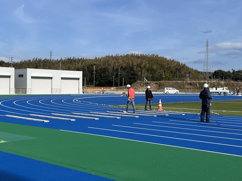 ヘルメットを被り防寒着を着用した3名の作業員が、カーブしたトラック部分のラインマーキング工事を行っている様子の写真