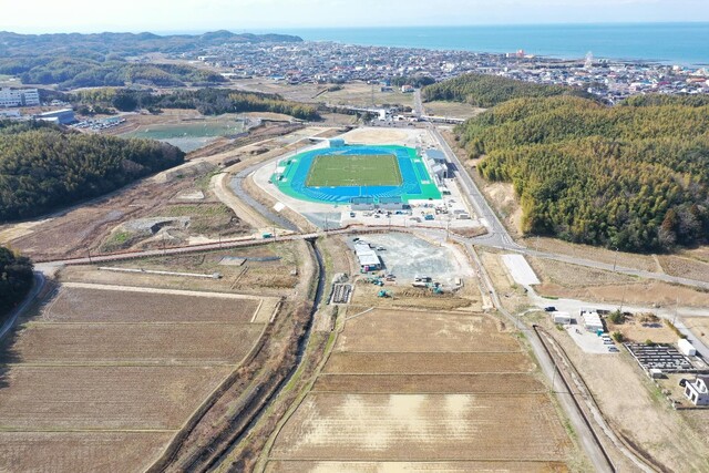 周囲に民家や鬱蒼と茂った森が点在する陸上競技場の建設予定地で工事が進められている様子を令和6(2024)年4月の全景を斜め上空から撮影した写真