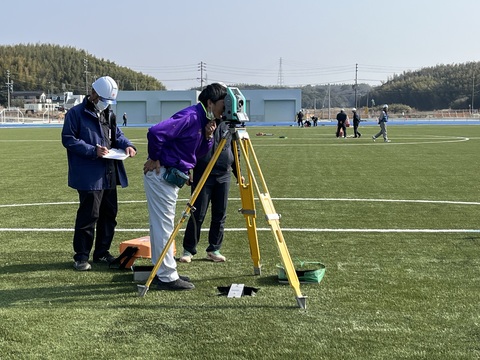 測量士が測量機器で検査を行い、後ろでヘルメットをかぶった作業員がメモを取っている写真