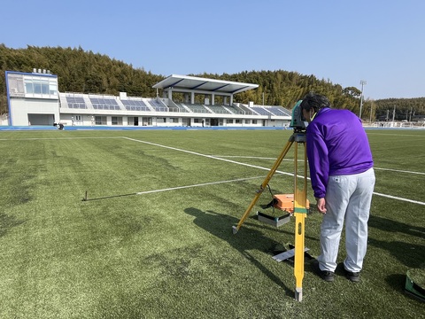 競技場内からスタンドの方向を見ながら測量士が測量機器で検査を行っている写真