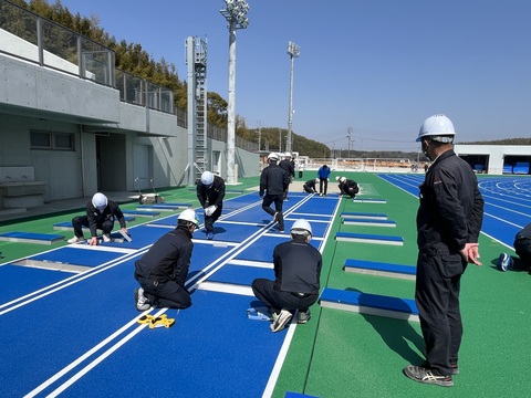 ヘルメットを被った作業員達が走り幅跳びコースのラインをチェックしている写真