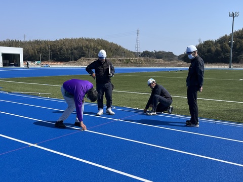 測量士の方が中腰でトラックのラインをチェックする様子をヘルメットを被った作業員達が見ている写真