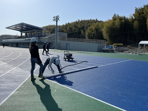 ヘルメットをかぶった作業員1人が右足でラインを押さえ、もう1人の作業員が機械を使い作業を行っている様子の写真