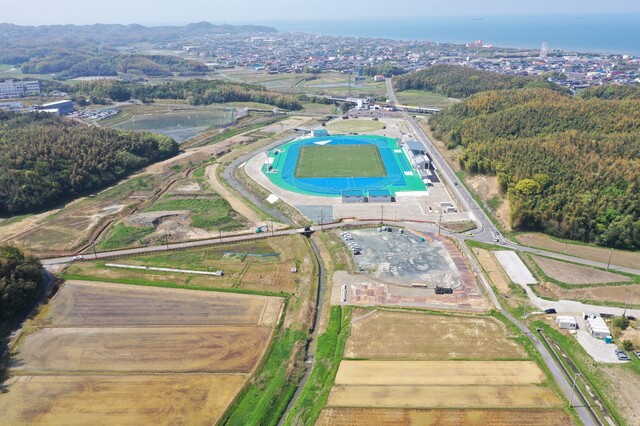 周囲に民家や鬱蒼と茂った森が点在する陸上競技場の建設予定地で工事が進められている令和6(2024)年4月の全景を斜め上空から撮影した写真