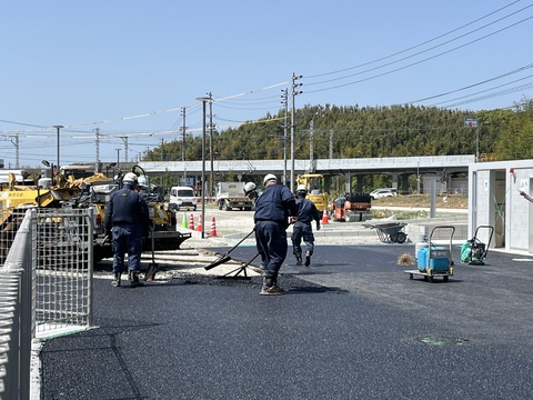 奥にコンバインドローラがあり、手前ではヘルメットを被った作業員達がレーキを使い作業を行っている様子の写真