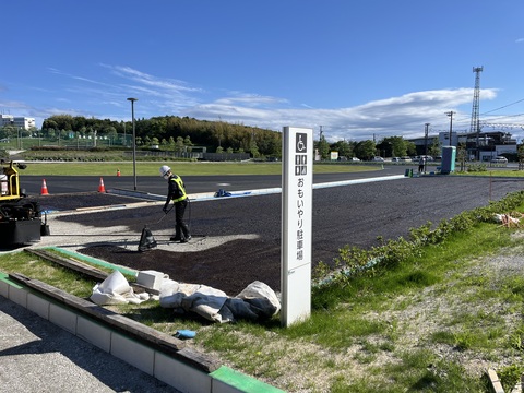ヘルメットを被り反射ベストを着た作業員が、「おもいやり駐車場」と書かれた看板が立っている奥の敷地でおもいやり駐車場の舗装工事を行っている写真