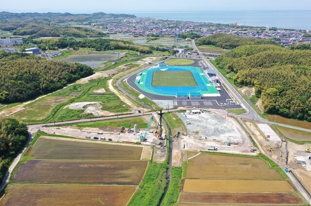 周囲に民家や鬱蒼と茂った森が点在する陸上競技場の建設予定地で工事が進められている様子を令和6(2024)年5月に斜め上空から撮影した写真