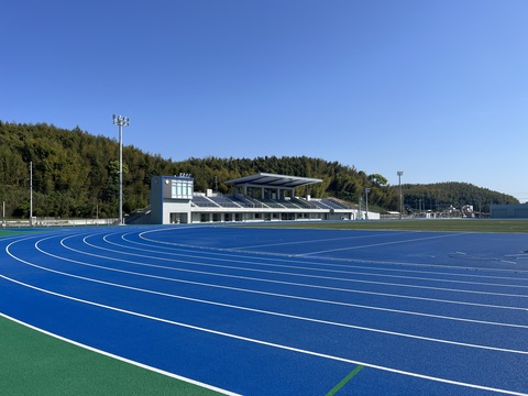 緑の木々を背景に、観覧スタンドと鮮やかな青いカーブしたトラック部分を撮影した完成写真