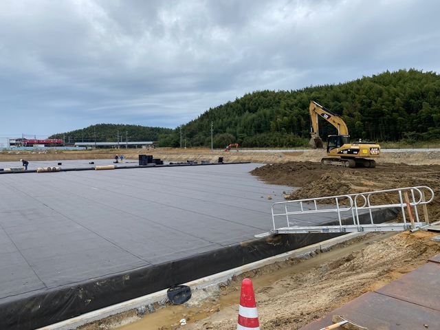 建設予定の陸上競技場に敷き詰められた黒色のプラスチック製の資材の上にシートが敷設され、その上に重機で土を被せている工事の様子写真