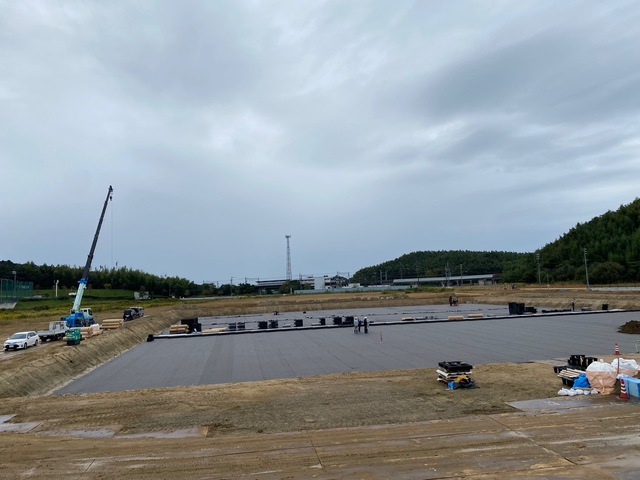建設予定の陸上競技場に敷き詰められた黒色のプラスチック製の資材の上にシートが敷設され、その周りの土手に資材が置かれ大きなクレーン車が停まっている工事の様子写真
