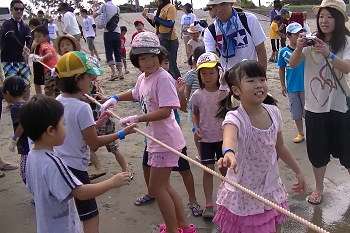 子どもたちを中心に砂浜から地引網を引っ張っている様子と、周りで多くの参加者たちが子供たちの様子を見守っている写真