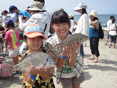 砂浜で、2人の女の子が両手で大きな魚をそれぞれ持ち一緒に記念撮影をしている写真