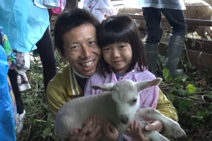 女の子と、女の子の後ろにいるお父さんが一緒に子ヤギを抱きかかえカメラに笑顔を見せている写真