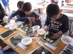 子供3人が横並びで、真剣な表情で出来上がった海苔にご飯をのせ恵方巻を作っている様子を写した写真