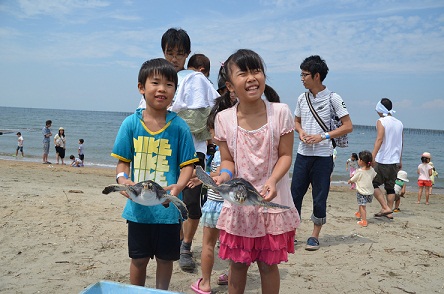 多くの人で賑わっている砂浜で、ウミガメを両手で持って笑顔を浮かべている子ども2人の写真