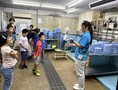 参加者たちが給餌室で、水色のシャツを着た飼育員の女性の話を聞いている様子の写真