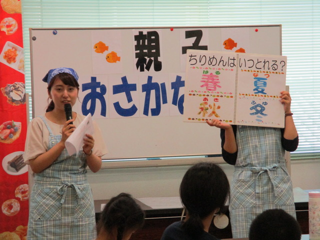 「ちりめんはいつとれる?春 夏 秋 冬」と問題の書かれたボードを持っている関係者、青い三角布をつけ、水色のエプロンを身に着けた女性がマイクを持って話をしている様子の写真