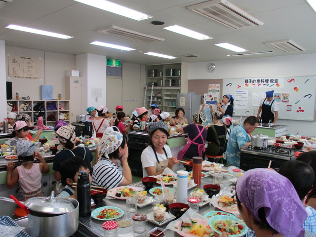 出来上がった彩りのある料理の品々が調理台に並び、参加者の親子が席について食事を楽しんでいる様子を写した写真