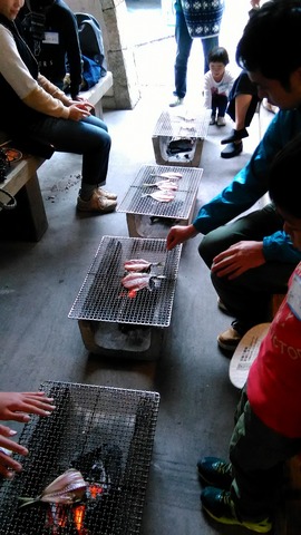 干物を網の上に並べて炭で焼きながら、周りに座って様子を見ている参加者達の写真