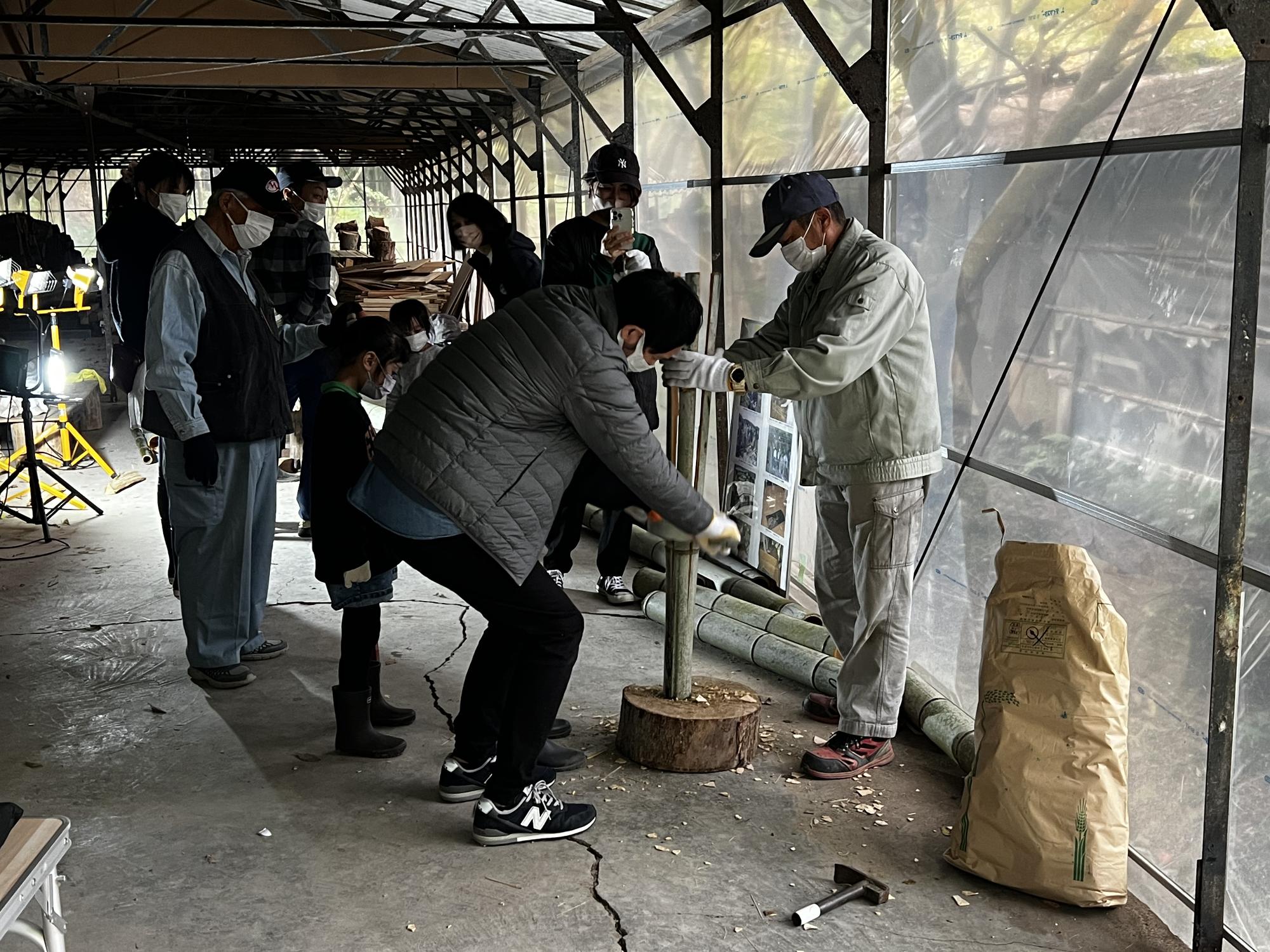 丸太の切り株の上に竹を立てて置き、一人の男性が竹を支え、もう一人の男性が竹を縦方向に割っている様子の写真