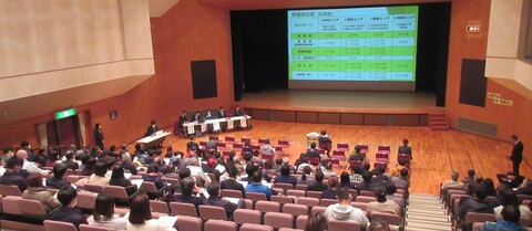 会場に多くの参加者が集まり、舞台上のスクリーンに資料が映し出されている説明会の様子を参加者の後方から写した写真