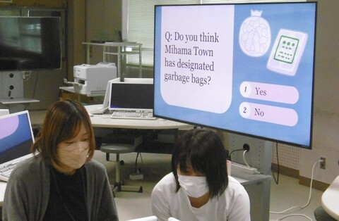 モニターに英語の問いと、「Yes」「No」の選択肢が表示されており、その手前でシェンさんが女子生徒に教えている様子の写真