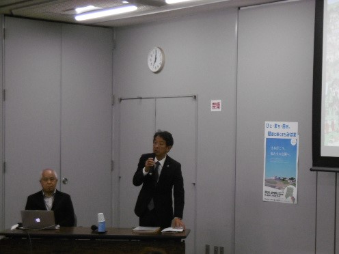 プロジェクタースクリーンの向かって左側に配置された席に2名の関係者がおり、右側の男性がマイクを手に立って話をしている写真