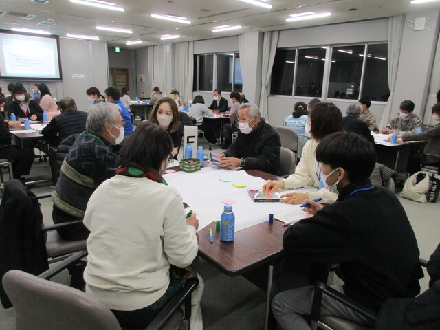 参加者がグループごとに分かれて席に着き、話し合いをしている会議室全体の様子を撮影した写真