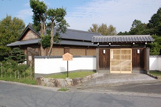 瓦屋根の門の先に、三角屋根に平屋建ての水野屋敷記念館が見える外観写真