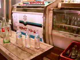 冷凍ショーケースの前に、水色や赤色のラベルが貼られた複数の飲み物の瓶が木製の台の上に置かれ展示されている写真