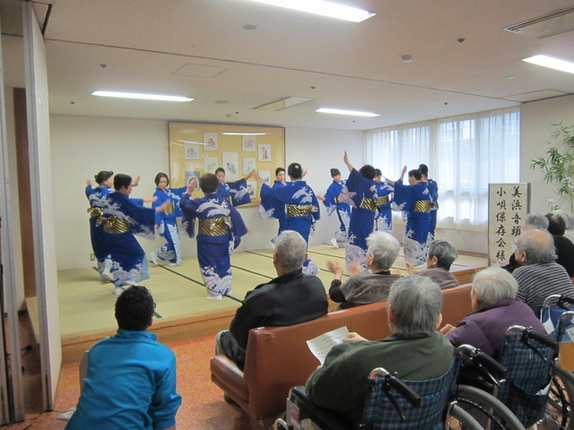 小上がりになった畳の上で、着物姿の関係者たちが輪を作って踊っている様子と、施設利用者たちが踊りを鑑賞している施設訪問の様子写真