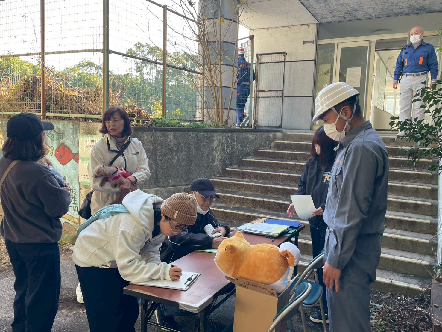 ペット避難訓練受付でアンケートに記入する2組の地元区住民。後ろで小型犬を抱えた2名の住民が談笑している。