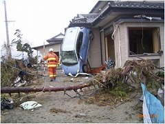 木は倒れ、窓がない家に、ブルーの車が逆立ちをしたように縦になり家に持たれかかった被災地の写真