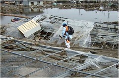 池のような水溜りが所々あり、ビニールハウスのようなものが倒れその上に箪笥がのっている間を隊員がヘルメットを被りリュックサックを背負い歩いている写真